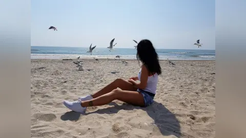 Young woman in a white tank top sitting on a sunny beach surrounded by flying seagulls.