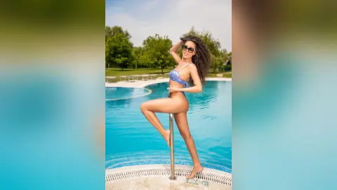 Smiling woman in a blue bikini posing by a sunny outdoor swimming pool on a bright summer day.