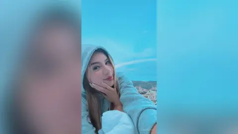 Smiling young woman in a cozy grey hoodie relaxing outdoors under a clear blue sky.