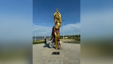 EnithAllegri poses outdoors next to a large golden statue of a woman under a clear blue sky.