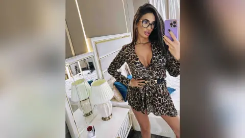AnnaKarev takes a mirror selfie wearing a low-cut leopard print dress and glasses while posing in her bedroom.