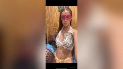 Young woman in a white ruffled crop top and pink masquerade mask posing in a cozy indoor setting.