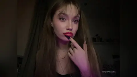 Young woman with long brown hair and red lipstick posing in moody, dim indoor lighting.