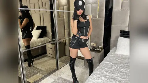 Woman in sexy black police officer costume and tall boots posing in a modern bedroom.