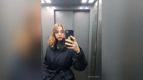Young woman with glasses in a black puffer jacket taking a mirror selfie in an elevator.