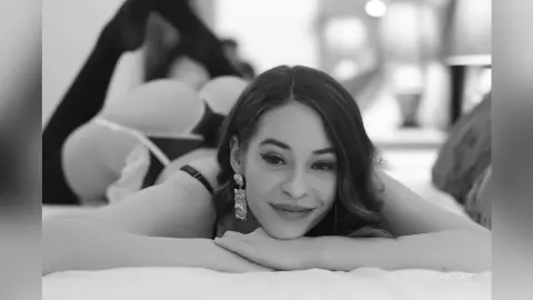 Smiling woman in black lingerie lying on a white bed in a soft, romantic black and white setting.