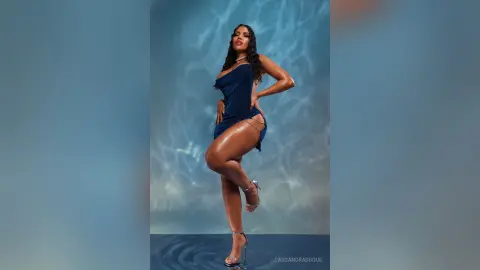 CassandraDuque poses in a short, tight blue dress and clear heels against a blue background, showing off her legs and curves.