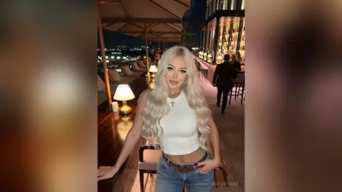 AlexaEvans poses in a white tank top and denim jeans, showing off her long blonde hair at an outdoor rooftop bar at night.