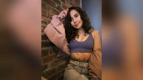 RossiNoleen poses against a brick wall wearing a blue crop top and light denim jeans.