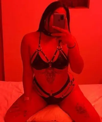 Woman in black strappy lingerie posing in a moody red lit bedroom setting.