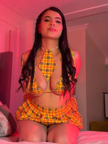 Young woman in a yellow plaid bikini set posing on a bed under warm pink lighting.