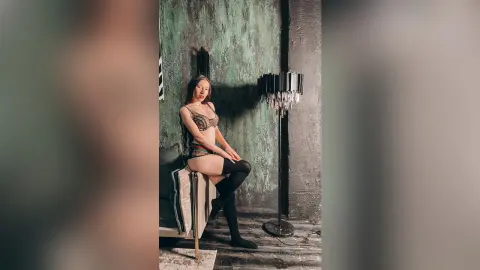 Young woman in patterned lingerie sitting on a chair against a moody, textured green wall.