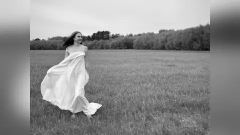 Smiling woman in a flowing white dress walking through a dreamy, misty meadow in a black and white landscape.