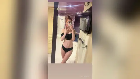 Young woman in a black bikini posing for a mirror selfie in a modern hotel room with warm lighting.
