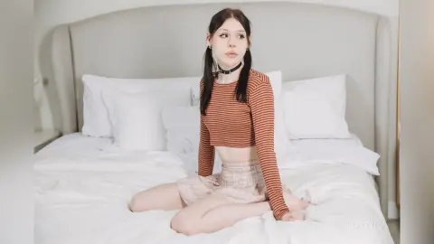 Young woman in a striped crop top and patterned skirt sitting on a white bed in a bright bedroom.