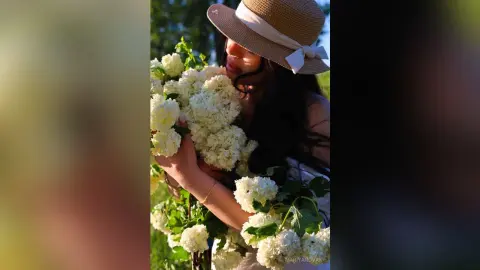 Dreamy portrait of a woman in a straw hat smelling white hydrangeas in a sunny summer garden.