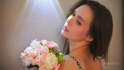 Dreamy portrait of a young woman with a pink flower bouquet against a textured wall in soft sunlight.