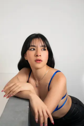 Susanmaeda with black hair and a blue strap top leans against a gray surface, looking away from the camera.