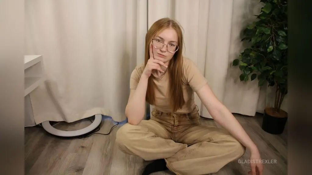 GladisTrexler — Redheaded woman in glasses and beige casual outfit sitting on floor in a bright room.