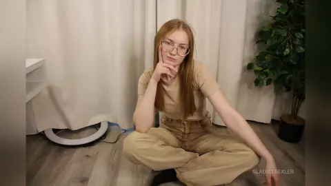 Redheaded woman in glasses and beige casual outfit sitting on floor in a bright room.