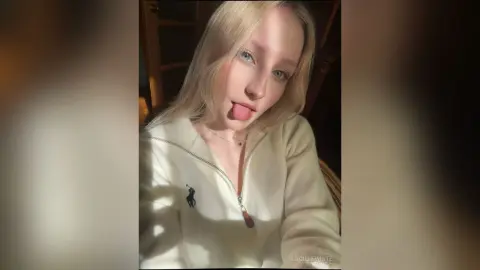 Blonde woman in a white zip-up sweatshirt making a playful expression in warm indoor lighting.