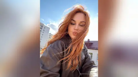 Redheaded woman in a black leather jacket blowing a kiss outdoors under a bright blue sky.