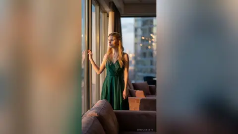Blonde woman in an elegant emerald green dress gazing out a high-rise window during golden hour.
