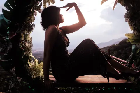 Raven_Morningstar sits in silhouette against a bright sky, wearing a dark dress and looking out over a mountain landscape.