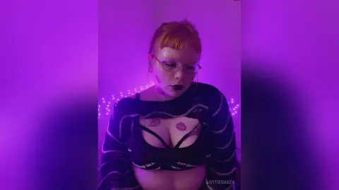 Redheaded woman in a black crop top and cardigan posing under moody purple neon lighting.