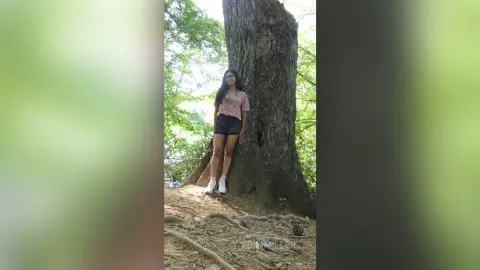 Smiling woman in a pink t-shirt and black shorts standing by a large tree in a lush green forest.