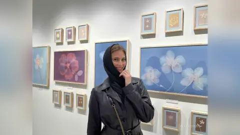 Smiling woman in a black leather jacket and hijab standing in an art gallery surrounded by floral paintings.