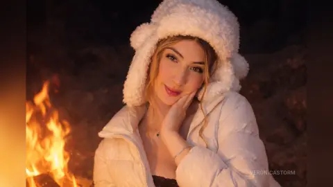 Smiling young woman in a white puffer jacket and fuzzy winter hat sitting by a warm campfire at night.