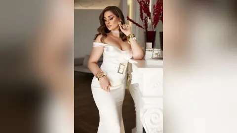 Elegant woman in a white satin evening gown posing glamorously in a bright, luxurious indoor setting.
