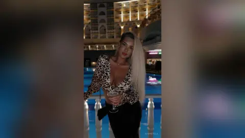 Glamorous woman in leopard print top holding a cocktail by a luxury resort pool at night.