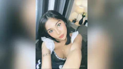 Young woman with long dark hair wearing a white ruffled black top in a cozy bedroom setting.
