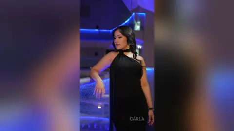 Elegant woman in a chic black halter dress posing in a modern luxury lounge with blue ambient lighting.