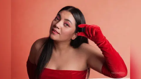 Young woman in a red strapless dress and matching velvet gloves posing against a warm pink background.