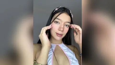 AleDilucas poses with her hands near her face, wearing a white and pink halter top with long black hair against a grey background.