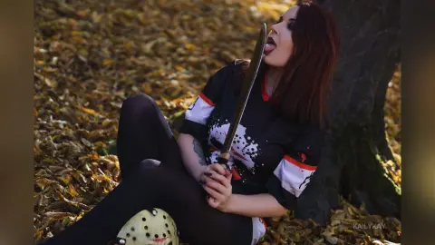 Woman in black t-shirt sitting in autumn leaves holding a prop sword with a playful expression.