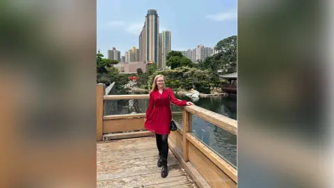 Smiling woman in a red dress standing on a wooden bridge overlooking a scenic city park and skyline.