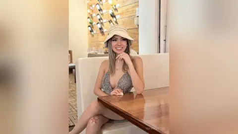 Smiling woman in a grey sequin top and beige bucket hat sitting at a wooden cafe table.