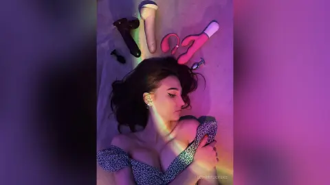 Young woman in a floral dress lying on a bed surrounded by adult toys under moody pink and green lighting.