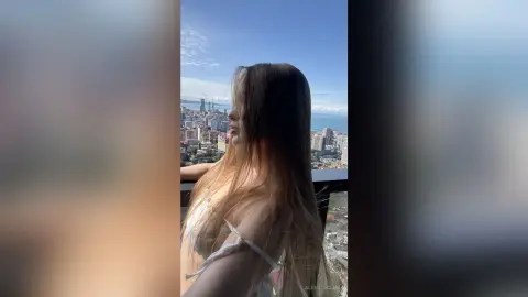 Long-haired woman in white lingerie looking at a city skyline from a high-rise balcony on a sunny day.