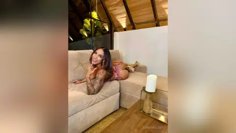 Smiling woman in pink lingerie relaxing on a beige sofa in a bright, modern sunroom setting.