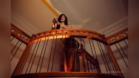 Elegant woman in a brown gown leaning on a wooden balcony railing in a warm, moody interior setting.