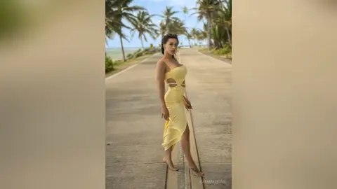 Elegant woman in a yellow cutout maxi dress walking on a tropical palm tree path during a sunny vacation.