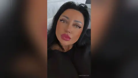 Glamorous woman with long dark hair and bold pink lipstick posing in a black top for a moody selfie.