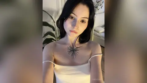 Young woman with dark hair and chest tattoo wearing a white camisole in a bright, minimalist room.