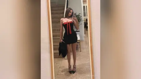 EvelynMills stands in front of a mirror wearing a short black strapless dress with a red ribbon detail.