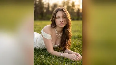 Young woman in a white lace dress and pearl necklace lying in a sunlit green field during golden hour.